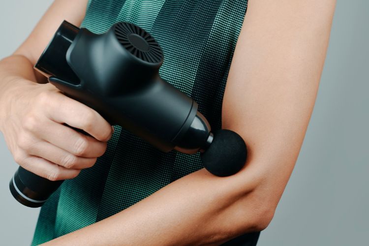Close-up van een persoon die een zwarte handmassagepistool op zijn bovenarm gebruikt. Het apparaat heeft een ronde, zachte bevestiging die tegen de huid wordt gedrukt. De persoon draagt een mouwloos, groen en zwart gepatroneerd topje. De achtergrond is neutraal grijs.
