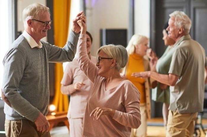 Twee glimlachende ouderen dansen samen binnenshuis, terwijl andere senioren op de achtergrond gezellig bij elkaar zitten en dansen, waardoor er een vrolijke, levendige sfeer ontstaat.