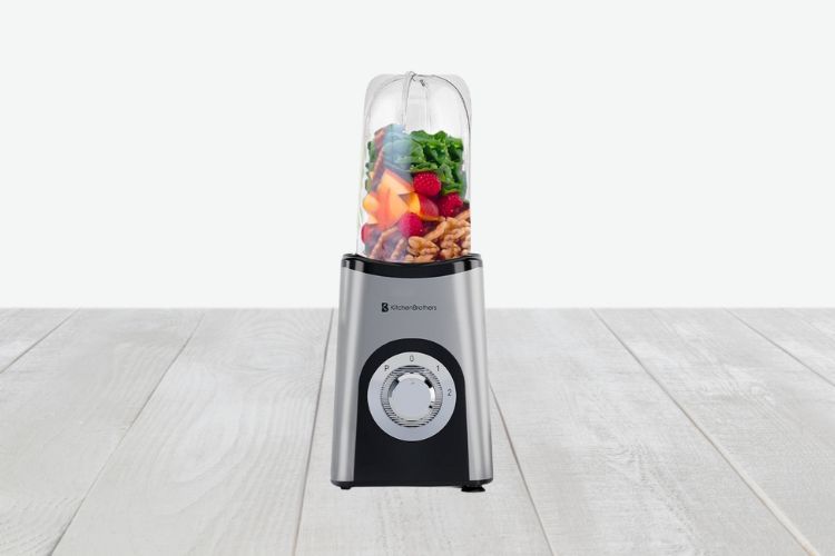 De KitchenBrothers Smoothie Blender, in strak zilver en zwart, heeft een transparante container gevuld met bladgroenten, wortels, rode bessen en walnoten. Met een ronde bedieningsknop met genummerde snelheidsinstellingen aan de voorkant, staat hij elegant op een licht houten oppervlak.