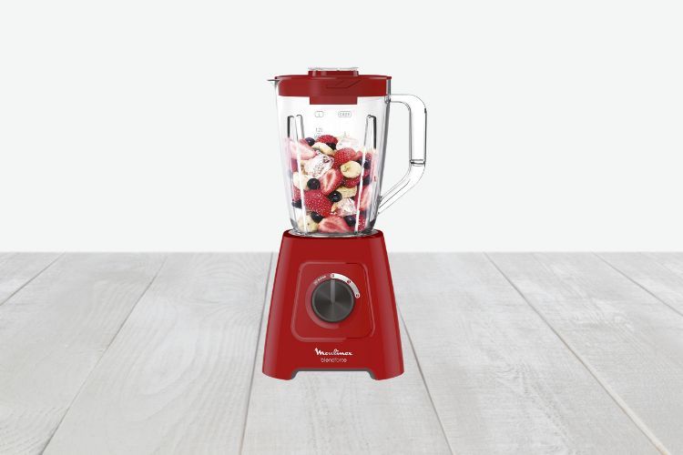 Een rode Moulinex blender met een draaiknop op de basis, met een mix van fruit, waaronder aardbeien, bosbessen en bananenschijfjes. Een van de beste smoothie mixers, staat op een licht houten oppervlak met een effen lichtgrijze achtergrond.