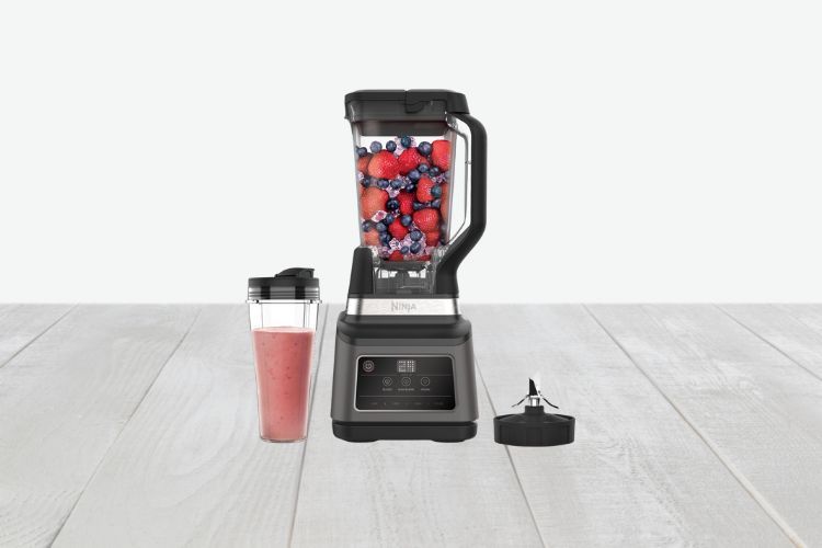 Een krachtige Ninja Foodi 2-in-1 Blender gevuld met gemengde bessen staat op een aanrecht. Ernaast staat een to-go beker met een roze smoothie en een deksel dat naast de blender rust. De scène is geplaatst op een licht houten oppervlak met een effen witte achtergrond.