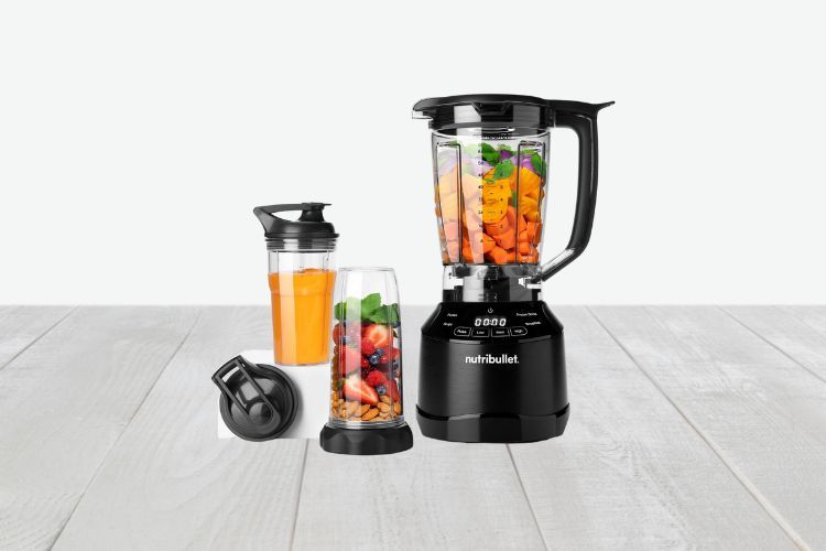 Een afbeelding van een Nutribullet Combo 1200 Watt Power Blender op een houten oppervlak. De blendercontainer is gevuld met gehakte groenten. Ernaast staat een reisbeker met een deksel en een andere afgesloten container gevuld met een oranje drankje. De achtergrond is een effen, lichte kleur.
