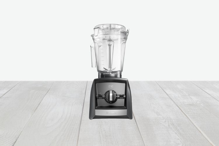 Een Vitamix A2500i Ascent-blender met een zwarte voet staat in het midden op een licht houten oppervlak tegen een effen witte achtergrond.