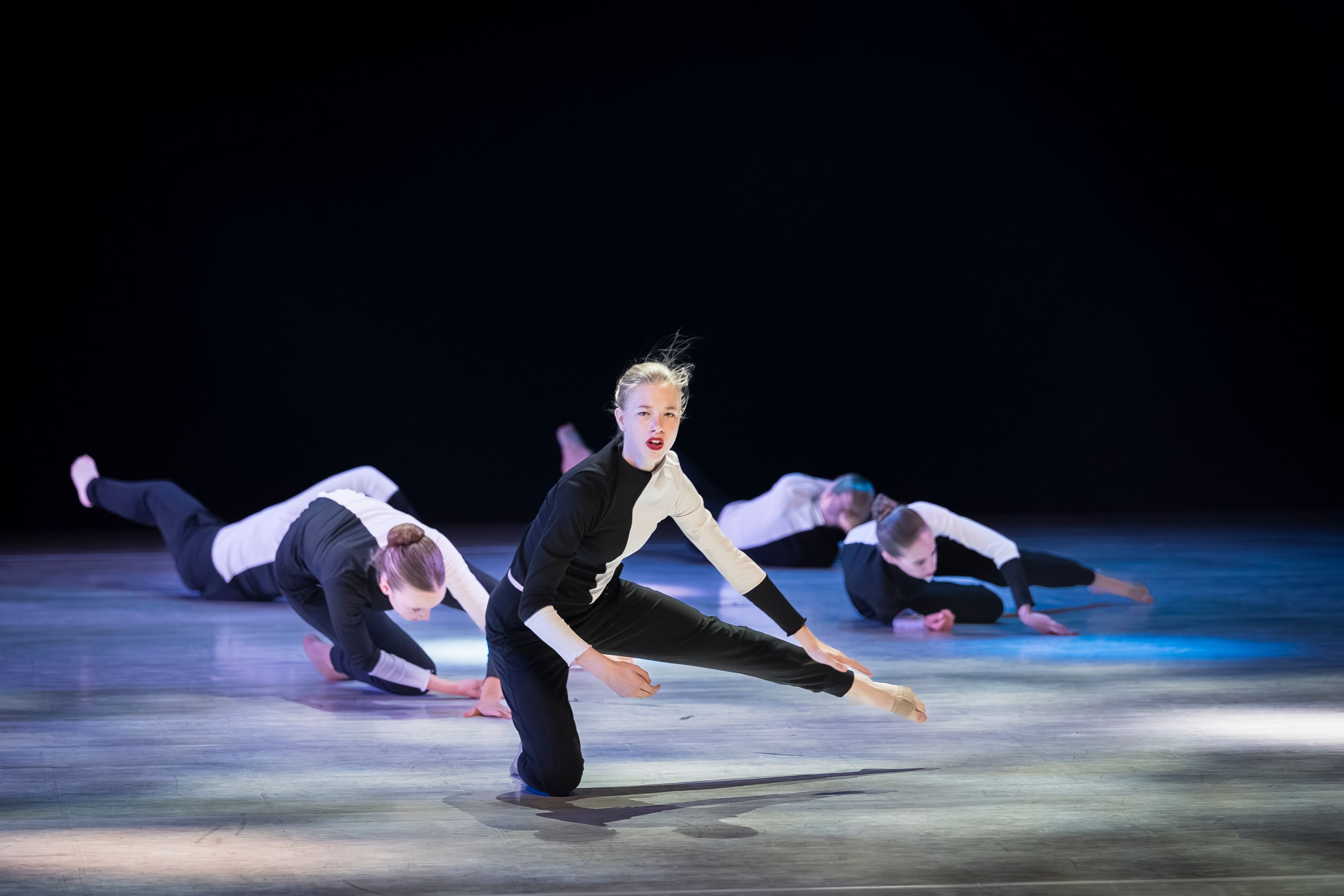 Een danser van Ndigo knielt op het donkere podium en kijkt recht in de zaal, terwijl de anderen gestrekt op de vloer liggen.
