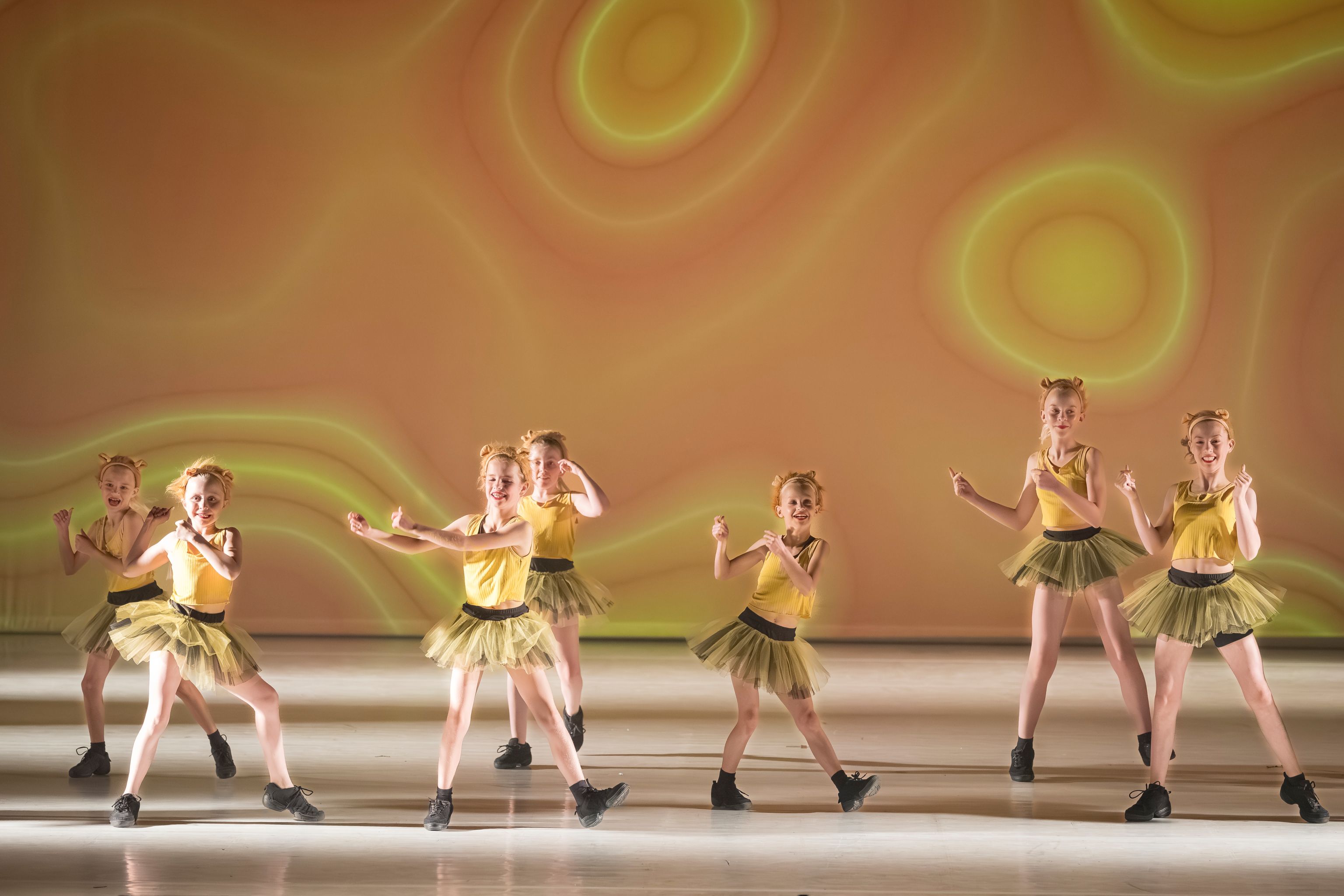 Creatieve dans voor kinderen bij Ndigo: zeven meisjes in gele kostuums en tule rokjes dansen synchroon op een sfeervol verlicht podium.