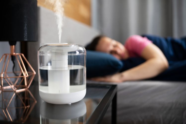 Een witte luchtbevochtiger stoot mist uit op een nachtkastje, vergezeld door een geometrische lamp. Op de achtergrond slaapt een persoon in een roze en blauw shirt vredig op het bed. De zacht verlichte slaapkamer is versierd met grijze gordijnen, wat een serene sfeer creëert.