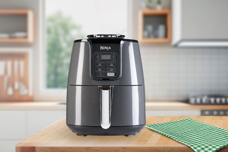 Een Ninja AF100EU-luchtfriteuse staat op een houten aanrecht met een groen geruit tafelkleed, met op de achtergrond een moderne keuken die vervaagt in zacht daglicht.