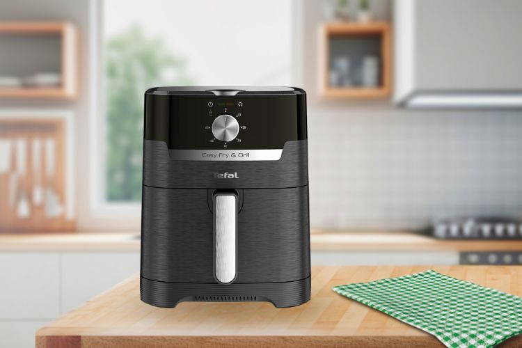 Een Tefal Easy Fry & Grill-heteluchtfriteuse staat op een houten aanrecht naast een groen geruit tafelkleed, met op de achtergrond een lichte, moderne keuken.
